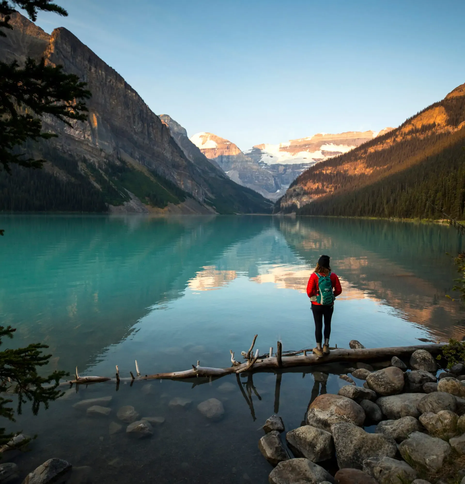 A hiker in a red jacket standing on a log at the edge of a calm, turquoise lake surrounded by steep mountains.