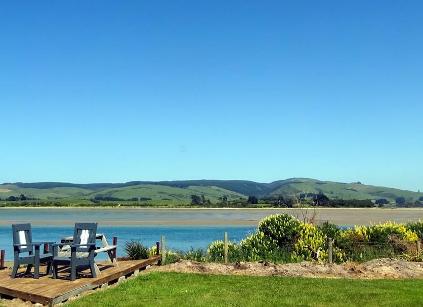 Surat Bay Lodge, Motel and Backpackers in Owaka with deck chairs by a river and hills beyond