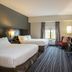The Mosey Buffalo Williamsville hotel room with two beds, desk, armchair and wall-mounted TV