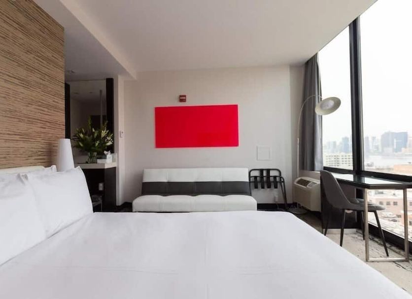 Z NYC Hotel Queens guest room with city view, floor-to-ceiling window, bed, sofa, and desk