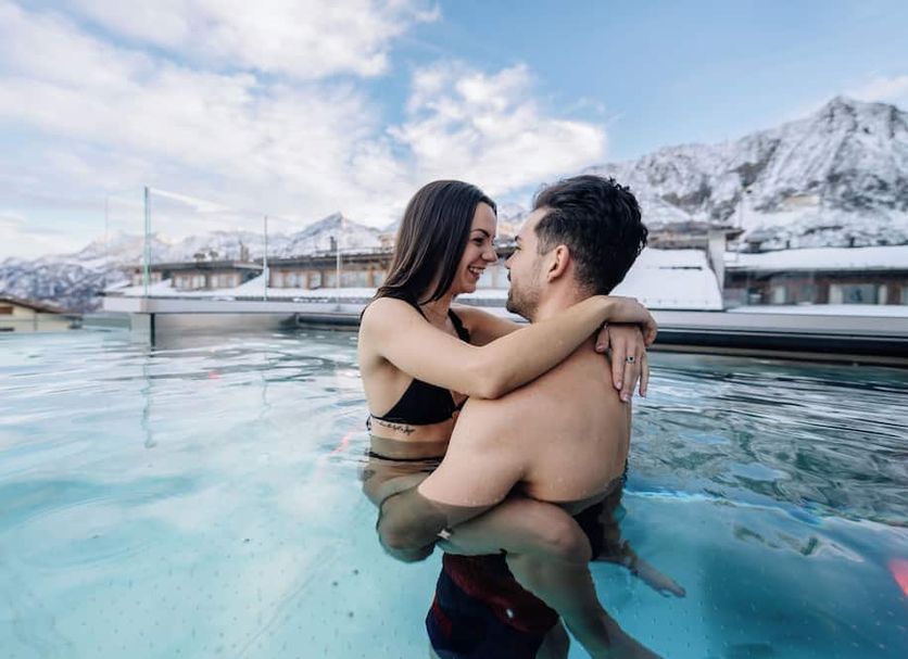 Couple embracing in an outdoor pool at Sporting Hotel, Passo del Tonale, with snowy mountains behind.