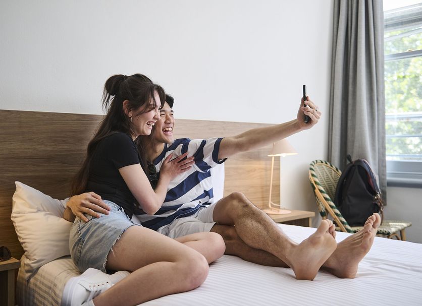 Guests taking a selfie on a bed at The Pacific House, Sydney hotel room with window light