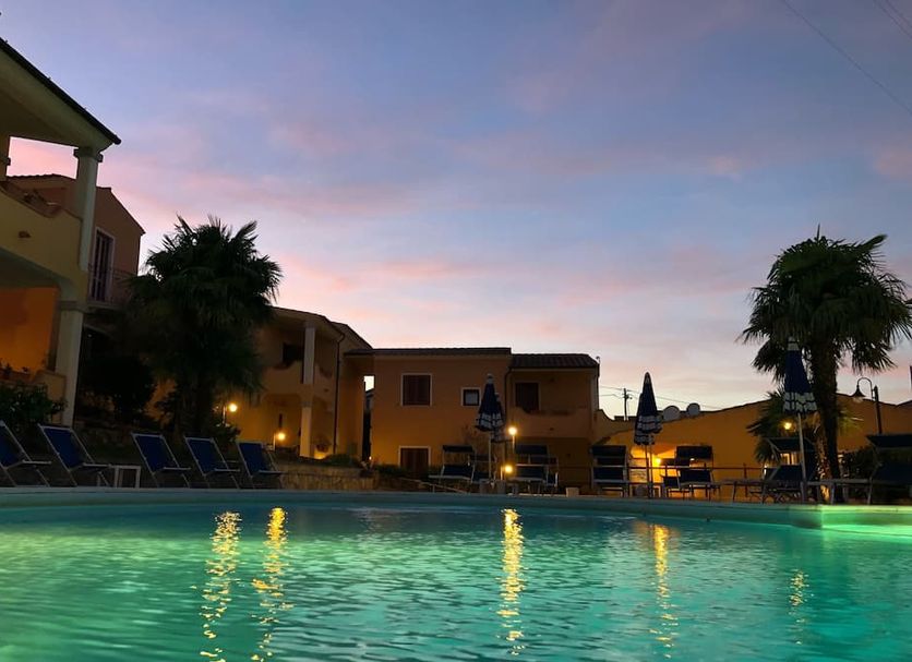 Residence Corte Dei Venti in Budoni with a lit outdoor pool at dusk and palm trees