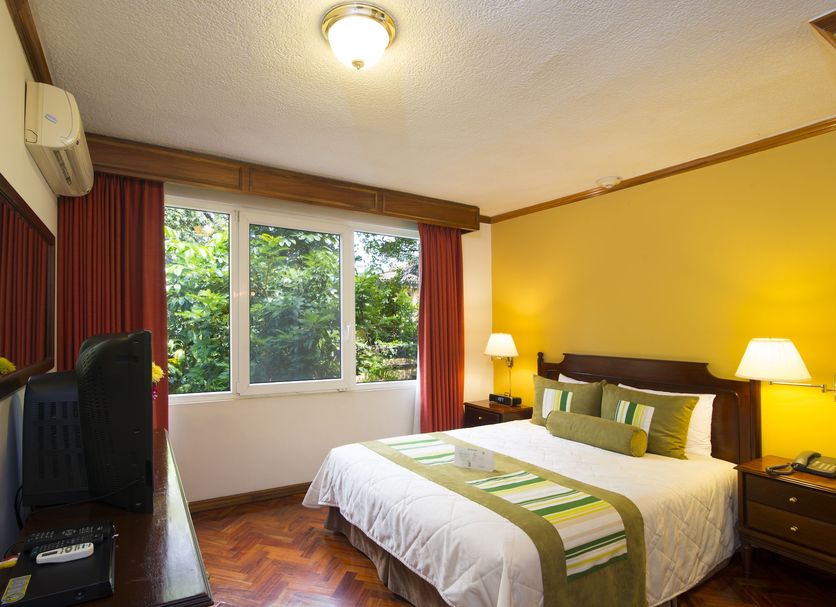 Apartotel & Suites Villas del Rio room in San José with a bed, desk, and window overlooking greenery