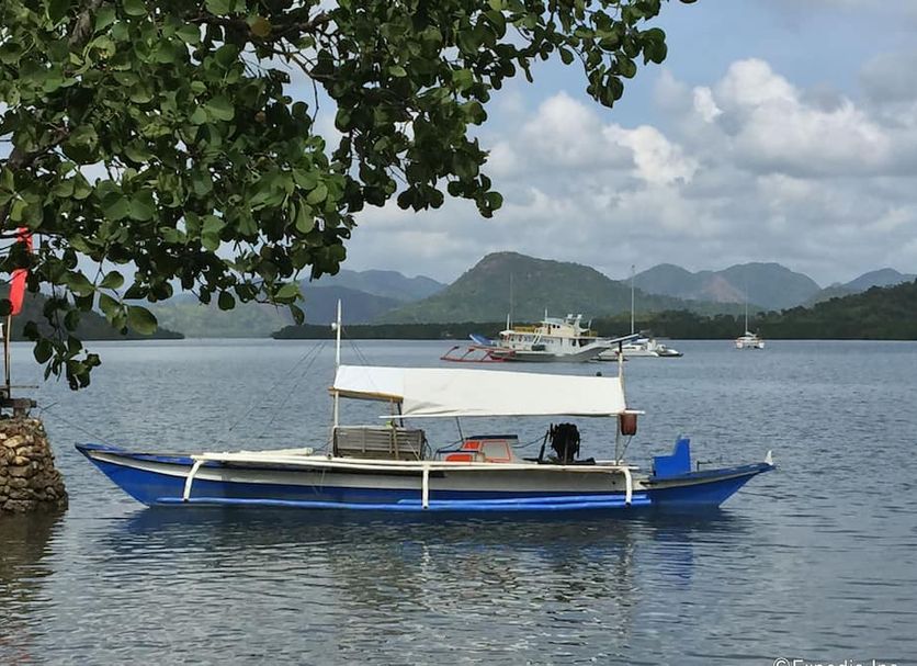 Discovery Island Resort in Coron: blue outrigger boat on calm bay with mountains and cloudy sky
