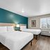 WoodSpring Suites San Antonio South guest room with two beds, teal accent wall, and window view of San Antonio