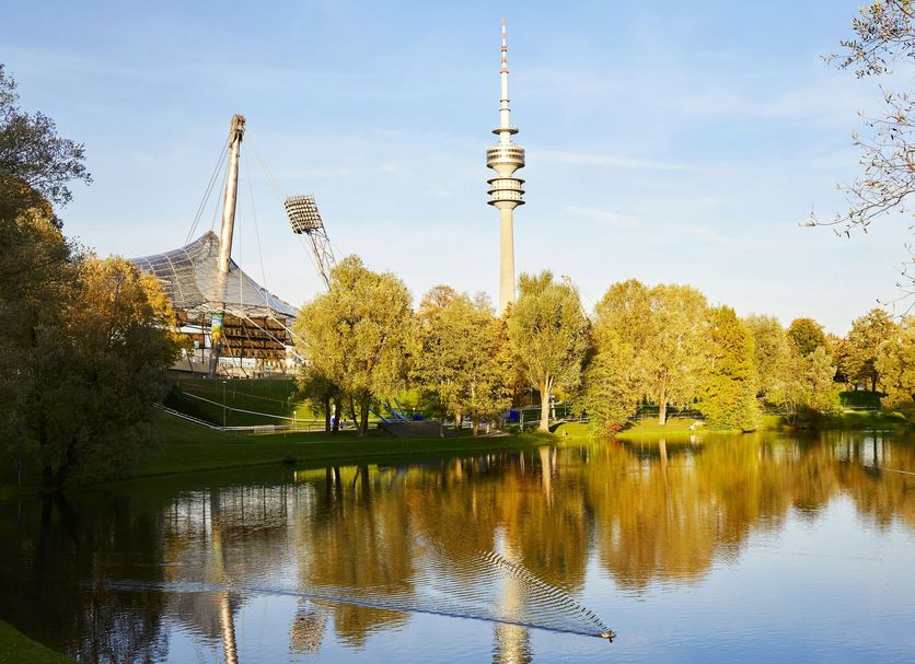 Living Hotel am Olympiapark in Munich with lake reflection, trees, and Olympiapark tower view