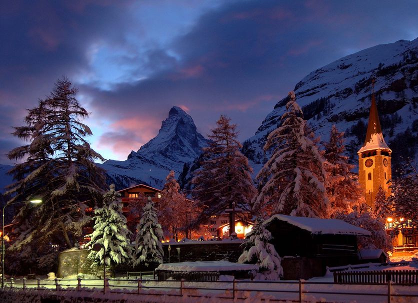 Swiss Alpine Hotel Allalin in Zermatt with snowy trees, illuminated church tower and mountain backdrop at dusk