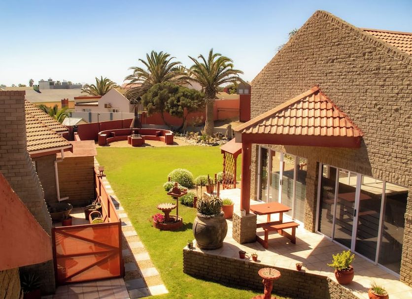 Amjicaja Guesthouse, Walvis Bay courtyard with palm trees, lawn, seating, and thatched-roof buildings