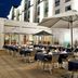 Outdoor terrace dining at Hilton Garden Inn Montreal Airport with tables, umbrellas and hotel facade.