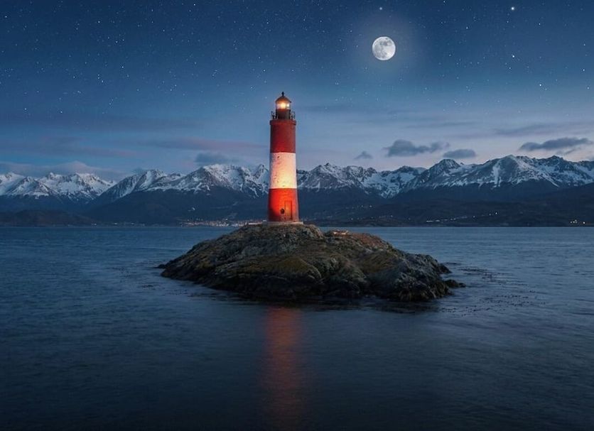 Hostal de la Laguna in Ushuaia with a lighthouse on a rocky islet beneath a moonlit mountain backdrop