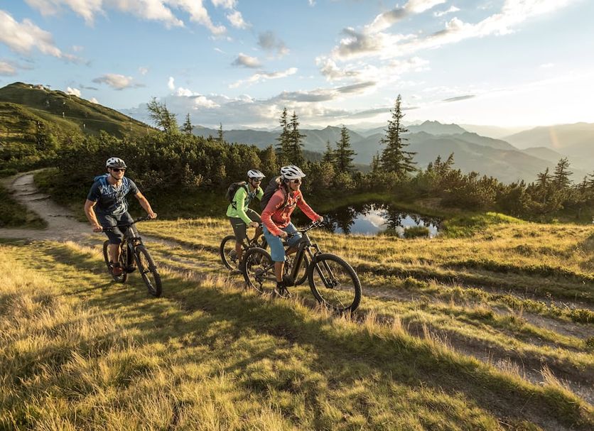 At Alpines Gourmet Hotel Montanara in Flachau, cyclists ride a grassy mountain trail at sunset.