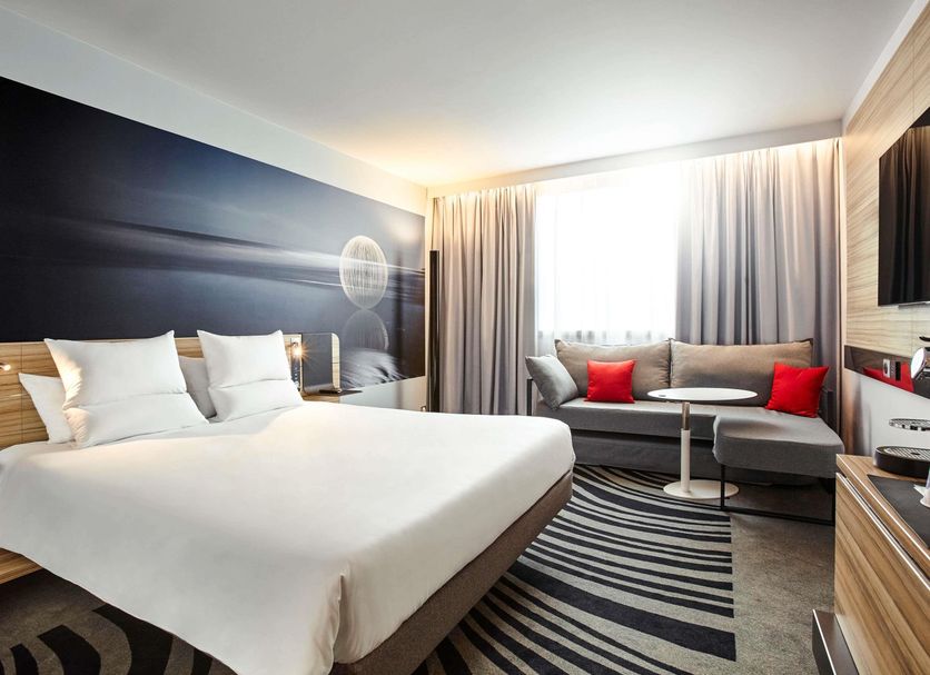 Novotel Narbonne Sud guest room with king bed, sofa seating, wall TV and striped rug in Narbonne