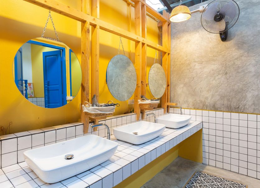 Aekkeko hostel in Phuket City bathroom with three sinks, round mirrors, yellow wood framing and wall fan