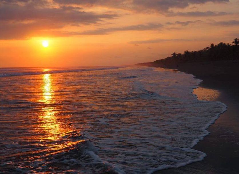 Sunset over the beach at Hotel Cocomar Beachfront Hotel in Parrita, with waves and palm-lined shore.