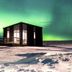 Afternoon Cottages in Hella, a small cabin on snowy ground beneath green northern lights