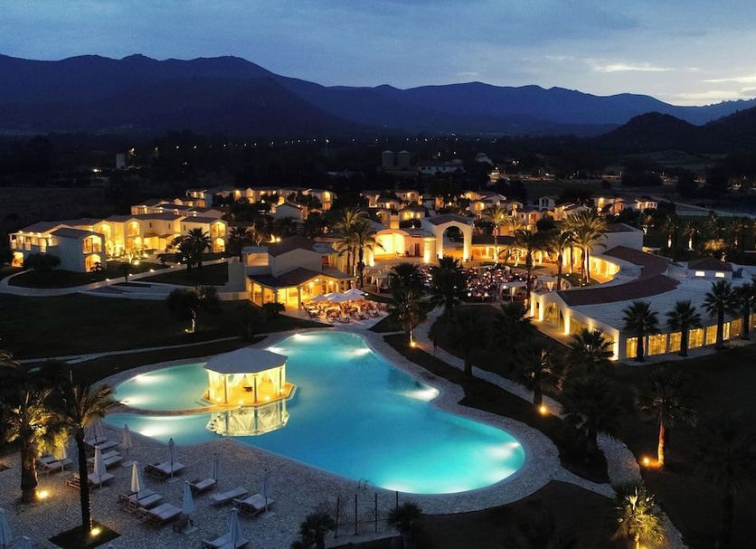 Spiagge San Pietro, Castiadas: illuminated resort pool and palm-lined grounds at dusk with mountain backdrop
