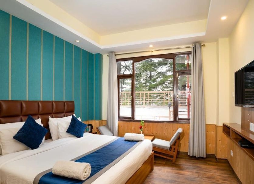 Hotel Woodrina in Shimla guest room with king bed, wall-mounted TV and window seat overlooking trees