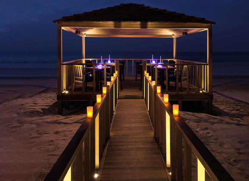 Dusit Ajman Resort & Villas in Ajman: lit wooden walkway to a beachfront pavilion at dusk