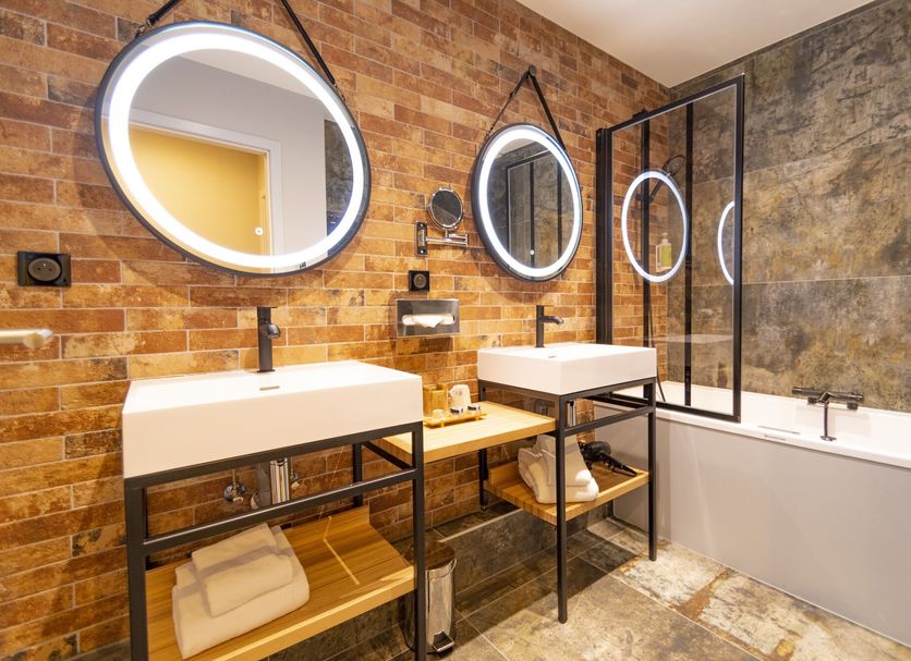 Hotel Beaucour, Strasbourg bathroom with double sinks, round mirrors, brick wall, and bathtub