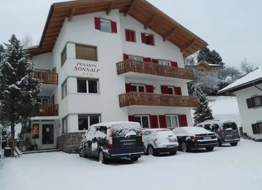 Pension Sonnalp in Ortisei, snowy alpine guesthouse exterior with balconies and parked cars