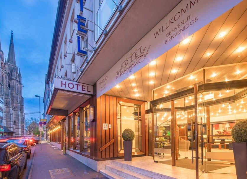 Hotel Marburger Hof entrance on a Marburg street at dusk, with lit glass lobby and cathedral nearby