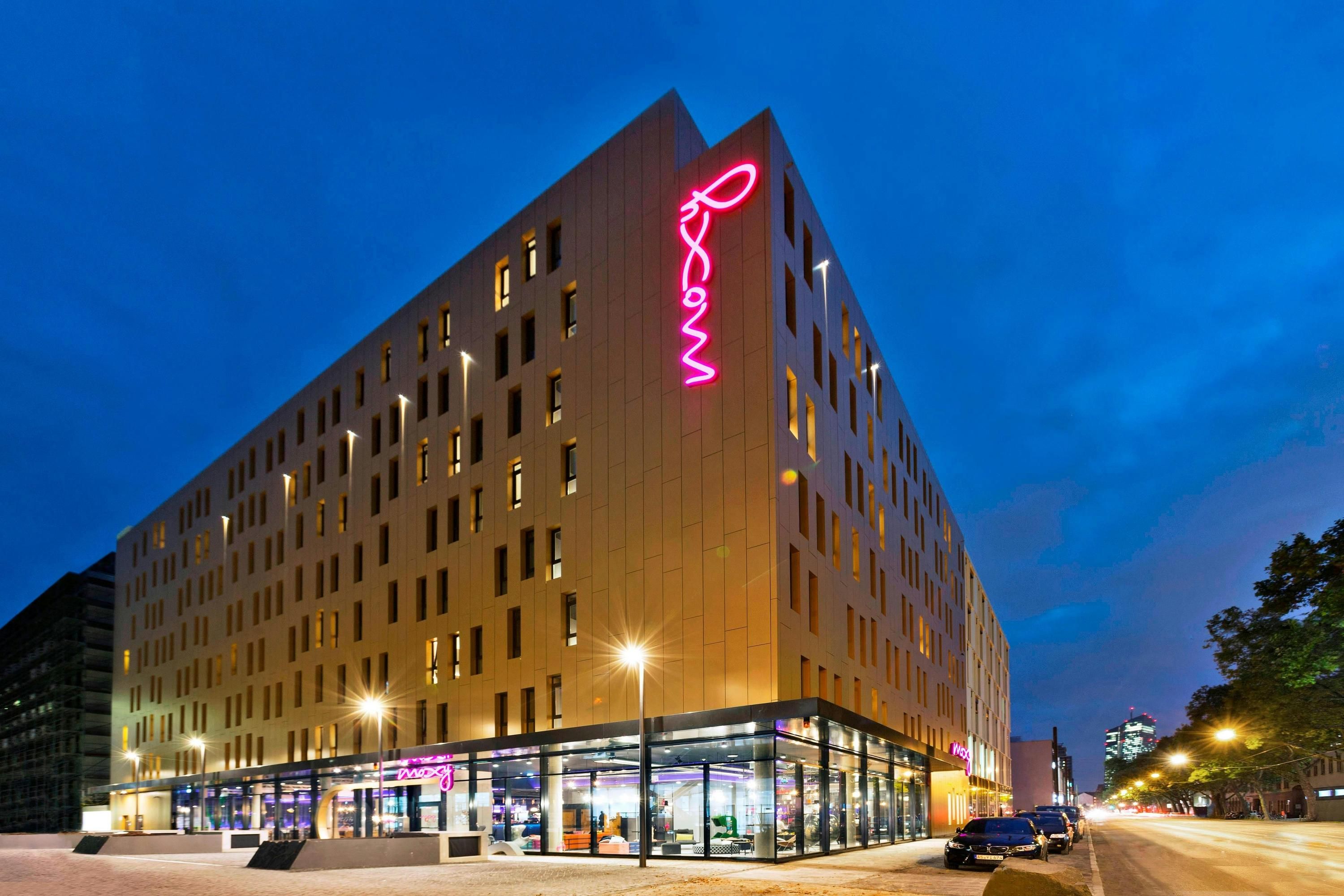 Moxy Frankfurt East in Frankfurt am Main, modern corner hotel exterior at dusk with neon sign