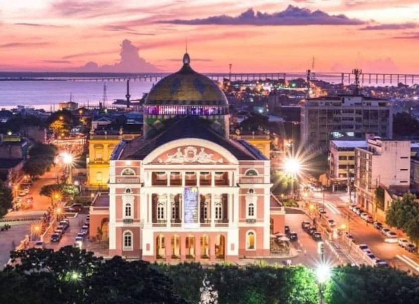 Hotel Dez De Julho in Manaus at sunset, with a domed historic building and river skyline beyond.