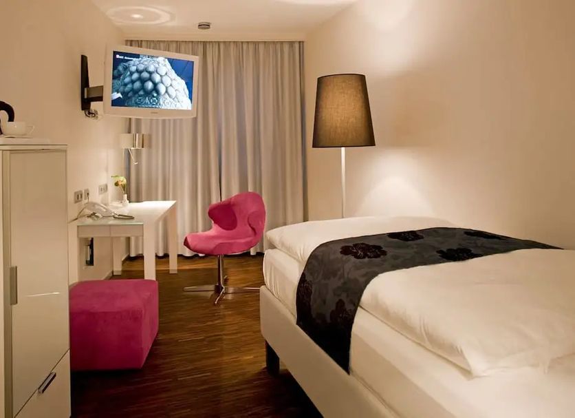 Zollamt Design Hotel Kaiserslautern guest room with bed, pink chair, desk, and window curtains