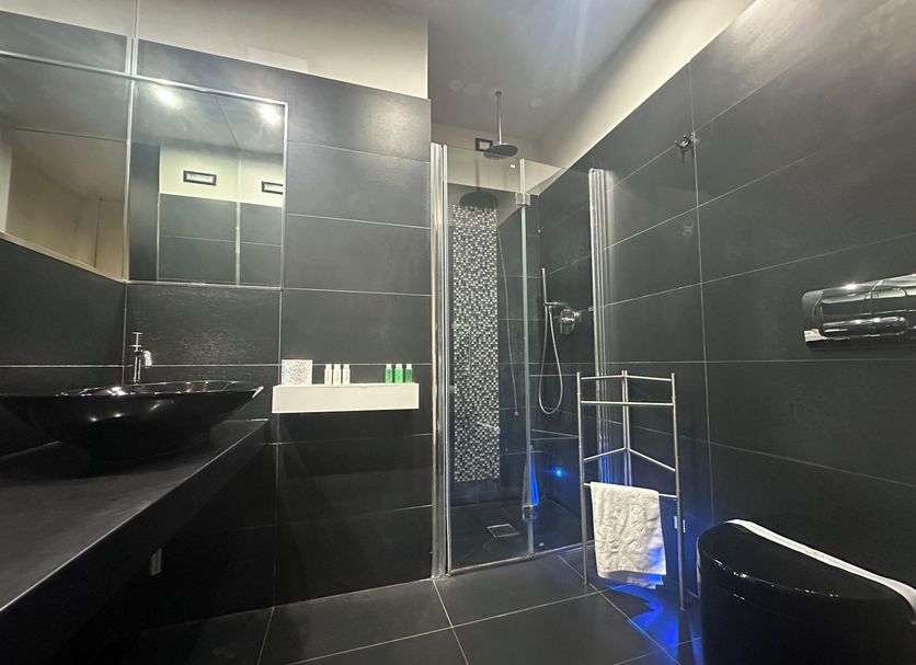 San Giorgio Modica Hotel bathroom with black tiled walls, glass shower, vanity sink, and blue floor lighting