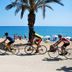 Cyclists ride past the beach at Hotel Alhambra in Santa Susanna, with palm tree and sea view.