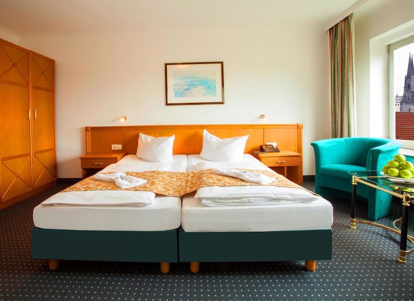 Ina Hotel Royal Cologne guest room with twin beds, teal chair, and city view through window