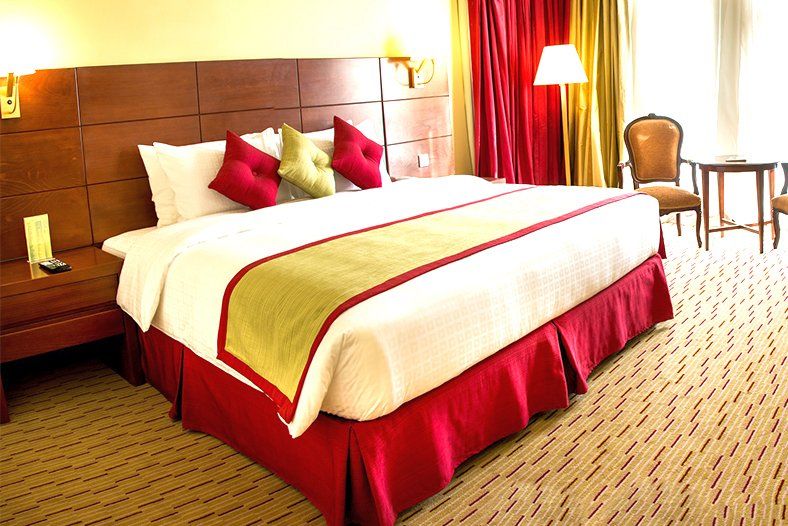 Rayan Hotel Sharjah in Sharjah guest room with a large bed, red accents, chair, and side table