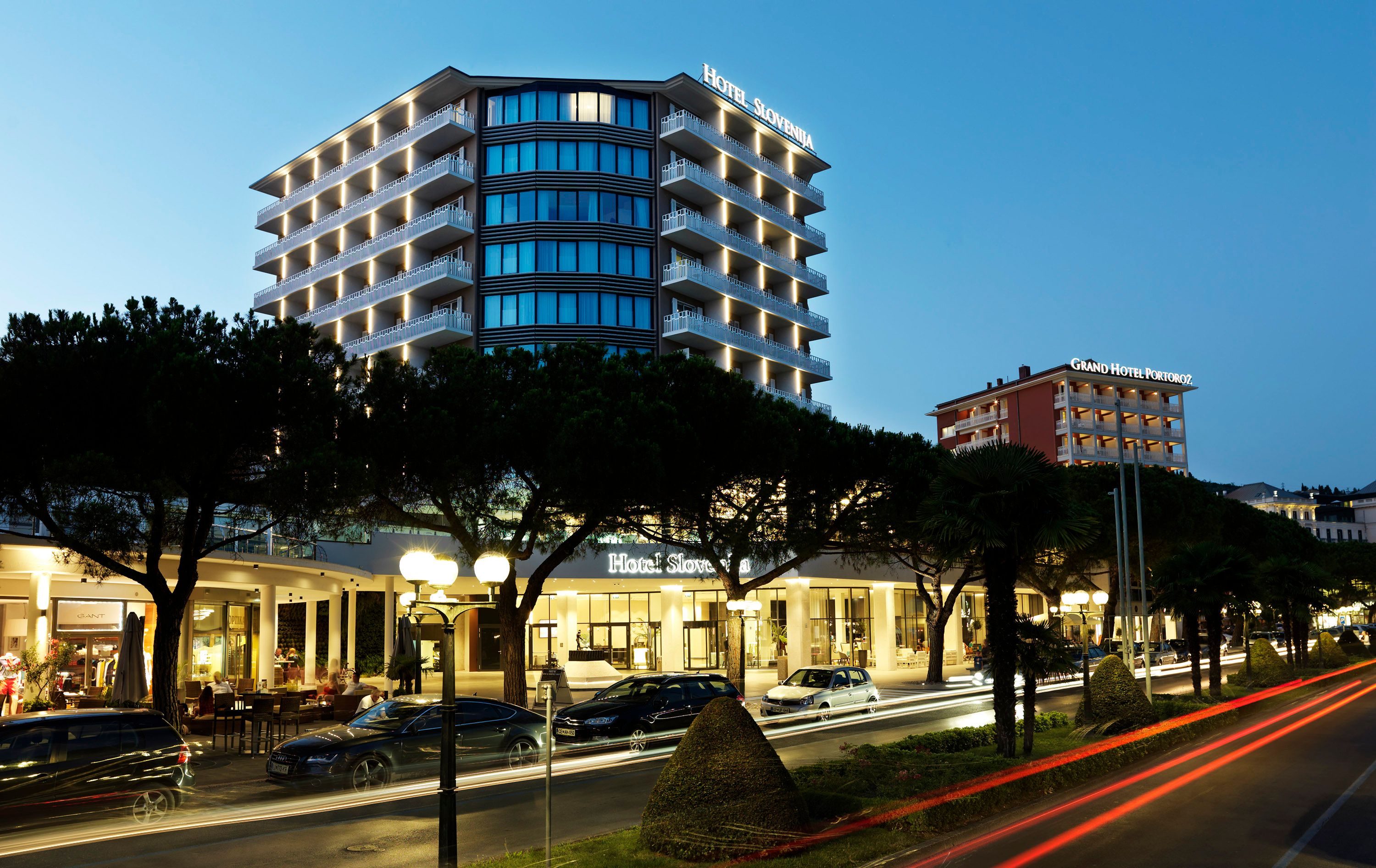 Mind Hotel Slovenija - LifeClass Hotels & Spa in Portorož at dusk, with seaside high-rise and lit street view.