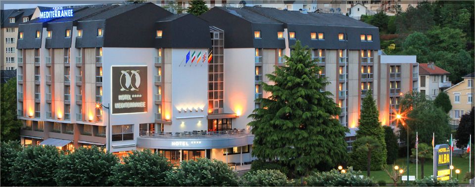 Hôtel Le Méditerranée in Lourdes, multi-story hotel exterior lit up at dusk with surrounding trees
