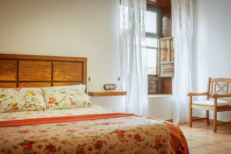 Casa Rural Arona guest room with floral bedspread, window, sheer curtains and wooden chair in Arona