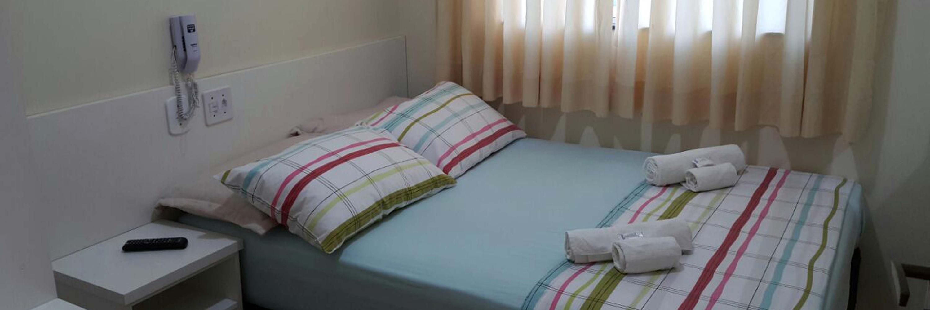 La Madre Hotel in Aparecida: simple guest room with double bed, striped pillows, towels, and curtained window.