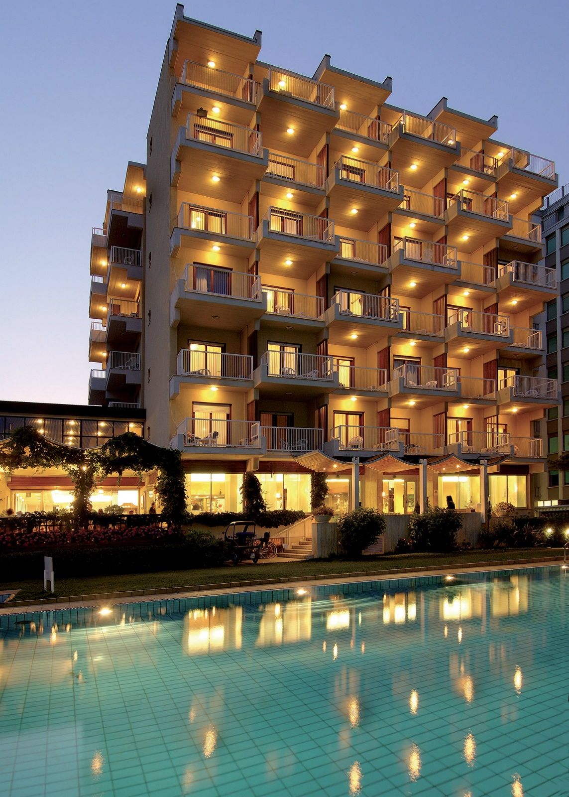 Hotel Atlantic in Lignano Sabbiadoro with lit balconies beside a reflective outdoor pool at dusk